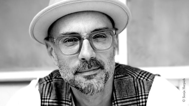 A photo of a middle-aged man with partially gray stubble and beard and large glasses with thick transparent frames. He is wearing a light-colored fedora hat and a plaid vest., © Sonja Herpich A photo of a middle-aged man with partially gray stubble and beard and large glasses with thick transparent frames. He is wearing a light-colored fedora hat and a plaid vest., © Sonja Herpich
