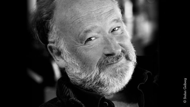 Black-and-white portrait photo of a smiling older male-identified person with a gray beard., © Stefan Gelberg Black-and-white portrait photo of a smiling older male-identified person with a gray beard., © Stefan Gelberg