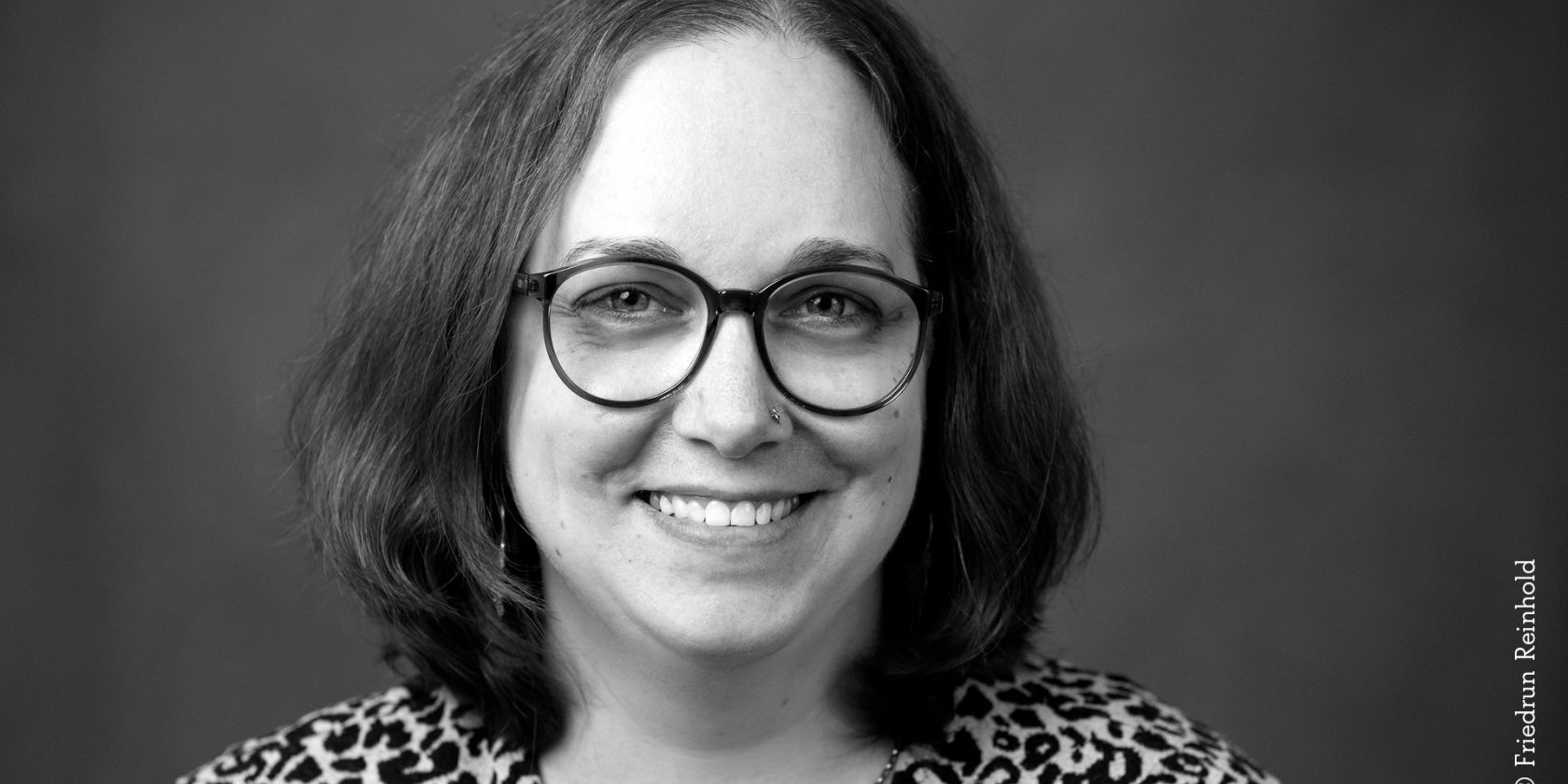 Black-and-white portrait photo of a woman with shoulder-length dark hair, a nostril piercing, and round glasses. She is smiling and wearing a shirt with a leopard print pattern., © Friedrun Reinhold Black-and-white portrait photo of a woman with shoulder-length dark hair, a nostril piercing, and round glasses. She is smiling and wearing a shirt with a leopard print pattern., © Friedrun Reinhold