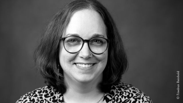 Black-and-white portrait photo of a woman with shoulder-length dark hair, a nostril piercing, and round glasses. She is smiling and wearing a shirt with a leopard print pattern., © Friedrun Reinhold Black-and-white portrait photo of a woman with shoulder-length dark hair, a nostril piercing, and round glasses. She is smiling and wearing a shirt with a leopard print pattern., © Friedrun Reinhold