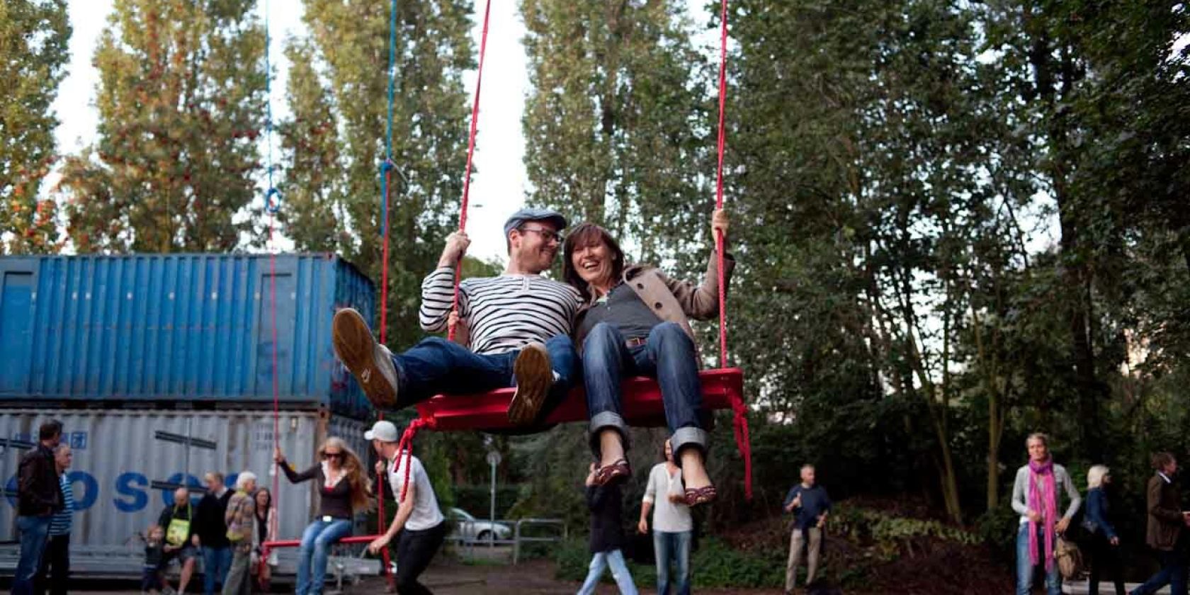 Zwei Menschen sitzen nebeneinander auf einer großen Schaukel im Kampnagel Garten und lachen. Im Hintergrund stehen einige andere Menschen vor Bäumen und Containern., © Antonia Zennaro Zwei Menschen sitzen nebeneinander auf einer großen Schaukel im Kampnagel Garten und lachen. Im Hintergrund stehen einige andere Menschen vor Bäumen und Containern., © Antonia Zennaro