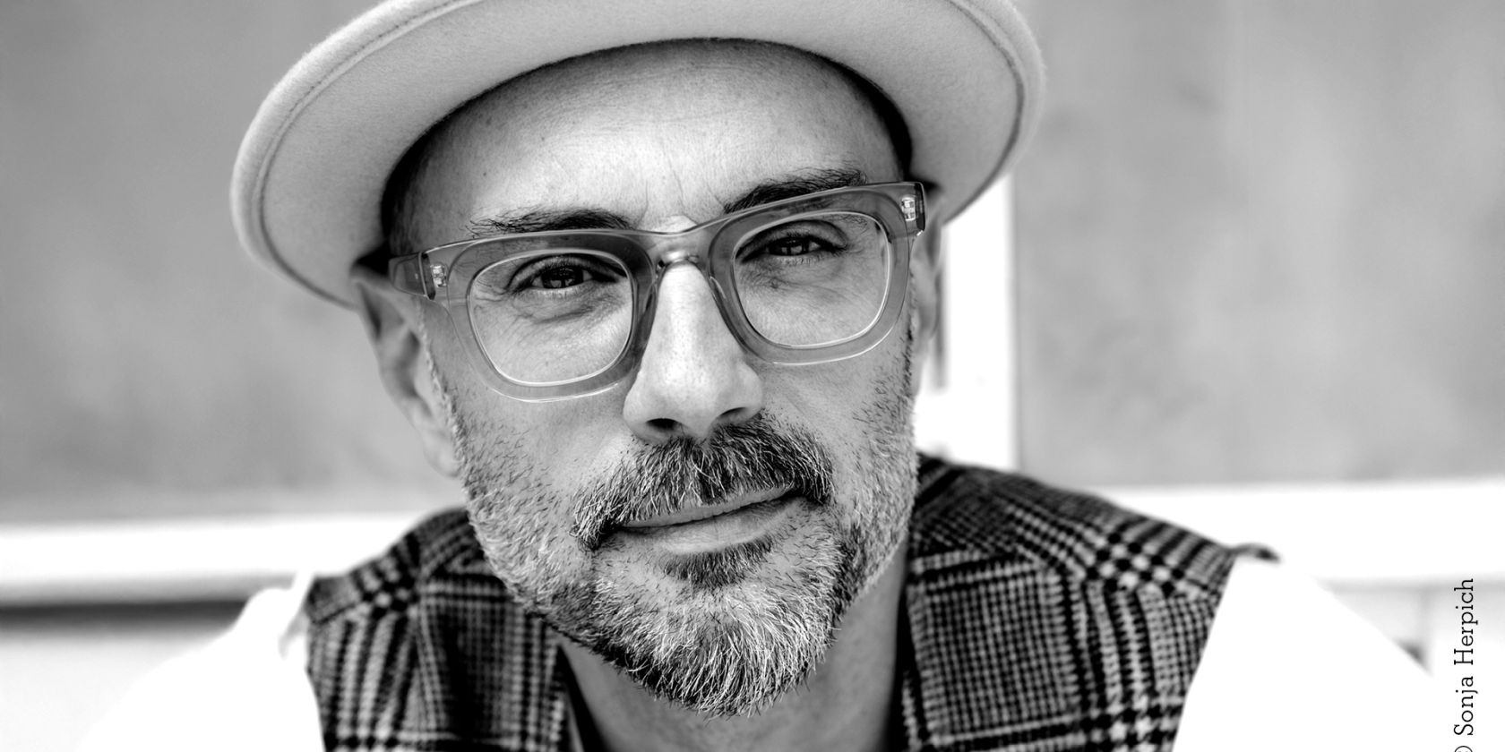 A photo of a middle-aged man with partially gray stubble and beard and large glasses with thick transparent frames. He is wearing a light-colored fedora hat and a plaid vest., © Sonja Herpich A photo of a middle-aged man with partially gray stubble and beard and large glasses with thick transparent frames. He is wearing a light-colored fedora hat and a plaid vest., © Sonja Herpich