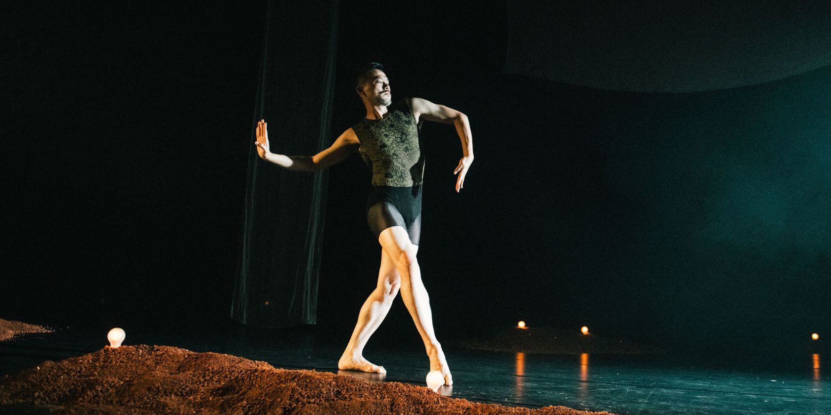 Ausschnitt aus der Tanzproduktion »Choreographing Story »Rama House«« von Chen Wu-kang und Pichet Klunchun. Eine performende Person bewegt sich in einer Ballett-Pose über die Bühne. Vor ihr ist ein kleiner erdartiger Hügel aufgescharrt., © Candy Weltz Ausschnitt aus der Tanzproduktion »Choreographing Story »Rama House«« von Chen Wu-kang und Pichet Klunchun. Eine performende Person bewegt sich in einer Ballett-Pose über die Bühne. Vor ihr ist ein kleiner erdartiger Hügel aufgescharrt., © Candy Weltz