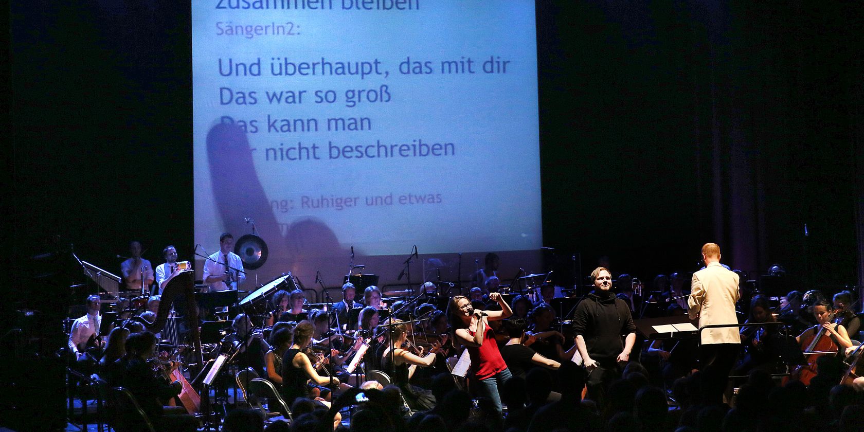 Ein Orchester spielt auf einer Bühne, im Hintergrund ist eine große Leinwand, auf die ein Songtext projiziert wird. Vor dem Orchester steht eine junge Frau, die fröhlich in ein Mikrofon singt., © Anja Beutler Ein Orchester spielt auf einer Bühne, im Hintergrund ist eine große Leinwand, auf die ein Songtext projiziert wird. Vor dem Orchester steht eine junge Frau, die fröhlich in ein Mikrofon singt., © Anja Beutler