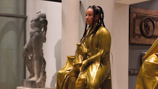 A person wearing a shiny gold-colored suit is on display as an exhibit in the museum., © Sarah Estio A person wearing a shiny gold-colored suit is on display as an exhibit in the museum., © Sarah Estio