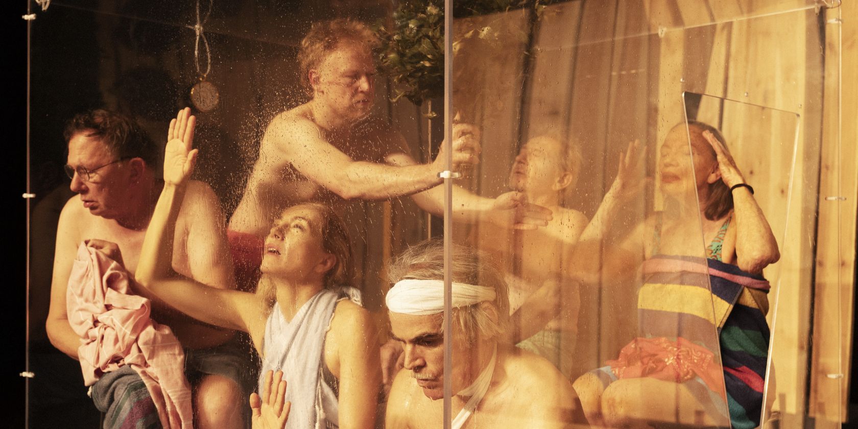 Sechs Performende sind in einer Sauna-Situation hinter einer Glaswand. Sie alle sind in unterschiedlichen Positionen und leicht bekleidet., © Armin Smailovic Sechs Performende sind in einer Sauna-Situation hinter einer Glaswand. Sie alle sind in unterschiedlichen Positionen und leicht bekleidet., © Armin Smailovic