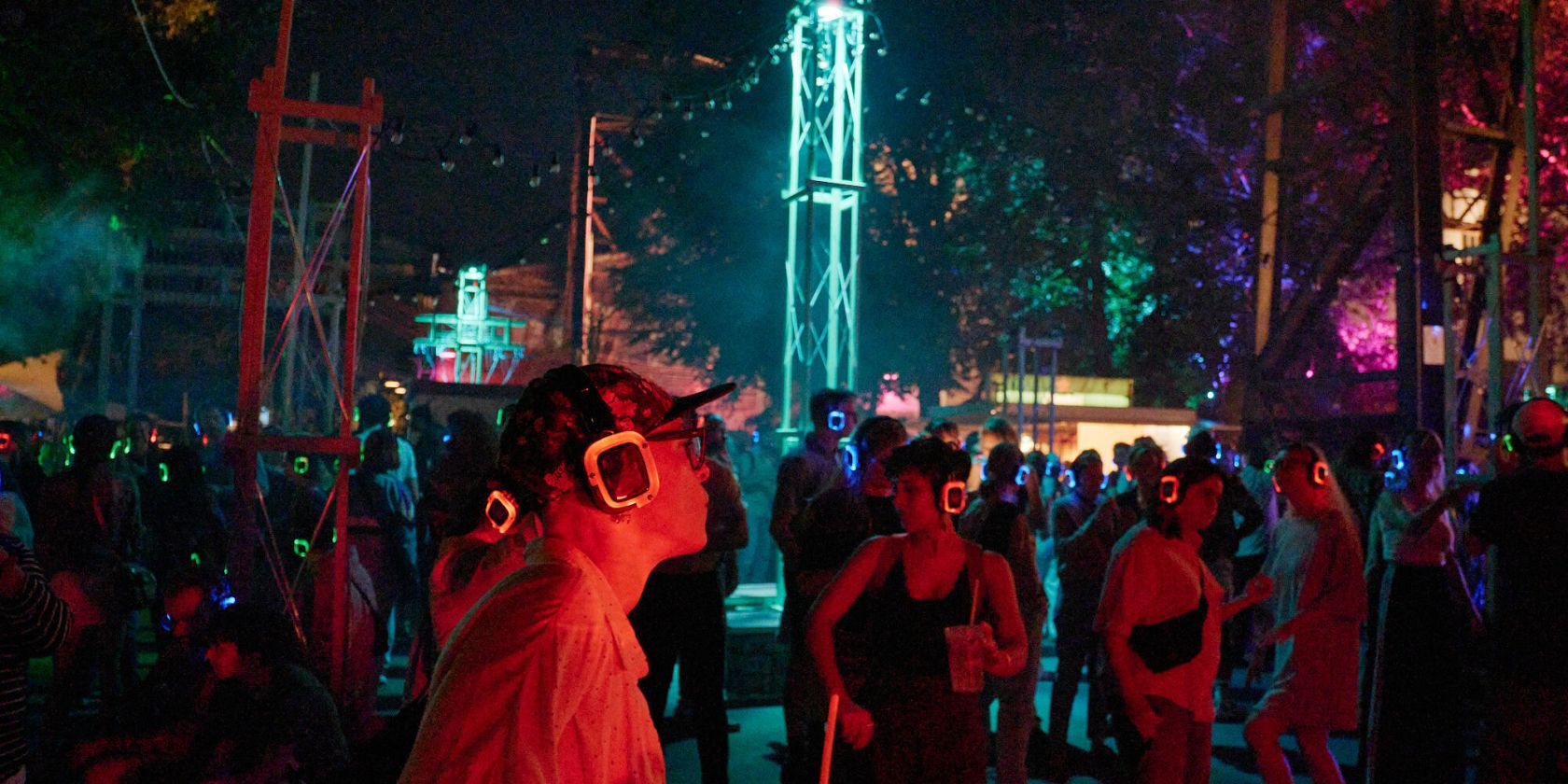 A nighttime JAJAJA event with a crowd wearing glowing red and blue silent disco headphones. In the foreground, a person wearing a cap and square glasses, holding a drink. In the background is an illuminated steel tower., © Fabian Hammerl A nighttime JAJAJA event with a crowd wearing glowing red and blue silent disco headphones. In the foreground, a person wearing a cap and square glasses, holding a drink. In the background is an illuminated steel tower., © Fabian Hammerl