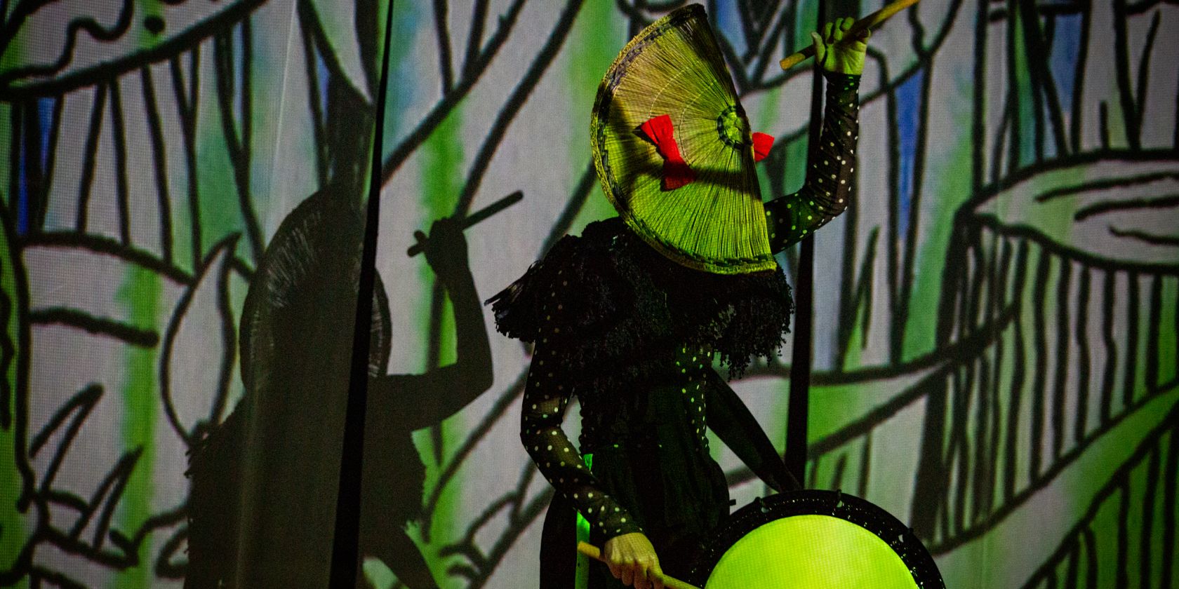 Eine Person trommelt vor einer in gr&uuml;n-schwarzen Symbolen angestrahlte Wand. Die eine Hand mit einem Drumstick in der H&ouml;he, die andere an der Trommel. Auf dem Kopf sitzt eine f&auml;cherartige Kopfbedeckung., &copy; Tsubasahori Maruszak