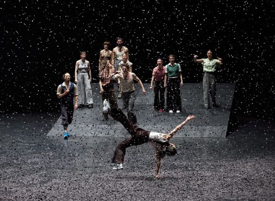 A group of dancers stands on a sloped stage while, in the foreground, one person balances impressively on one hand as confetti swirls through the air., © Dominik Mentzos