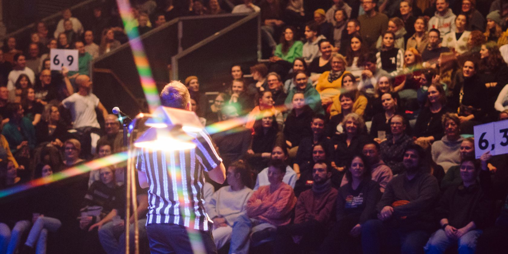 A person wearing a black and white striped T-shirt stands in front of a large audience. Two boards displaying scores are held up in the audience., © Michelle Jekel