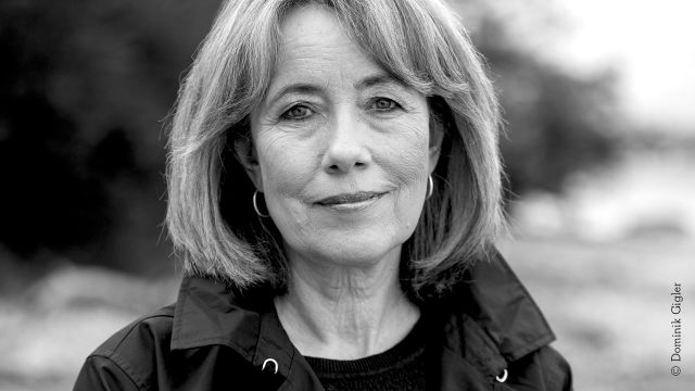 Black-and-white portrait photo of a middle-aged woman with a bob hairstyle. She is wearing hoop earrings and a dark jacket., © Dominik Gigler