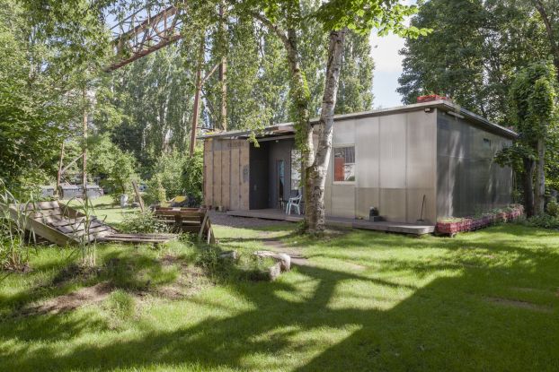 Photo of the Migrantpolitan at Kampnagel. A small one-storey house with a green garden, benches made of wooden pallets, surrounded by many trees., © L. Volkmann