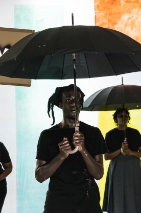 Ausschnitt aus der Produktion DAMBUDZO von nora chipaumire. Eine Person steht mit einem Regenschirm vor einer bunten Wand., &copy; Marie Staggat