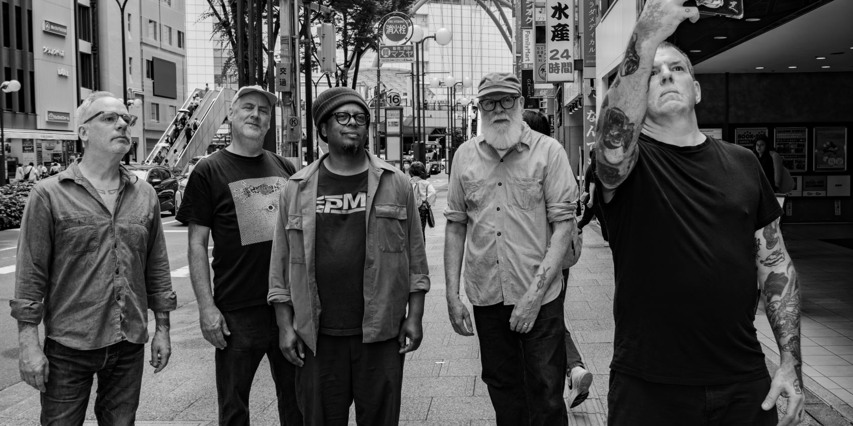 Five middle-aged male-looking people are standing together and looking up. One person with tattooed arms is holding up a cell phone and appears to be taking a selfie of the group. They are standing in the middle of a large Asian city., &copy; Yusuke Nagata