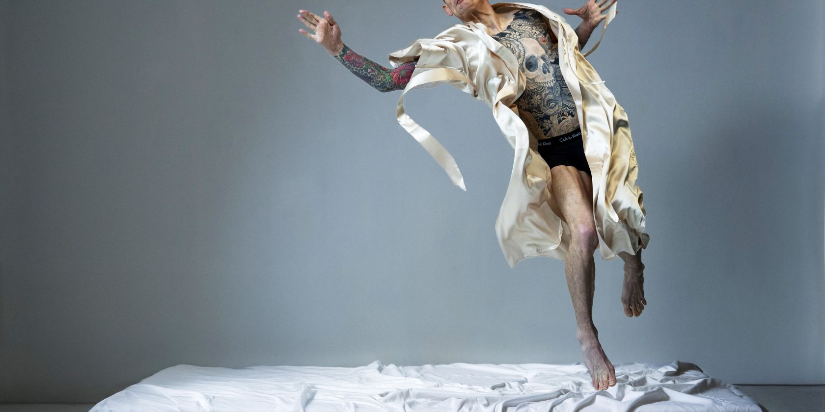A performer in a cream silk robe and black underwear leaps from a white bed, his torso intricately tattooed., © Hugo Glendinning