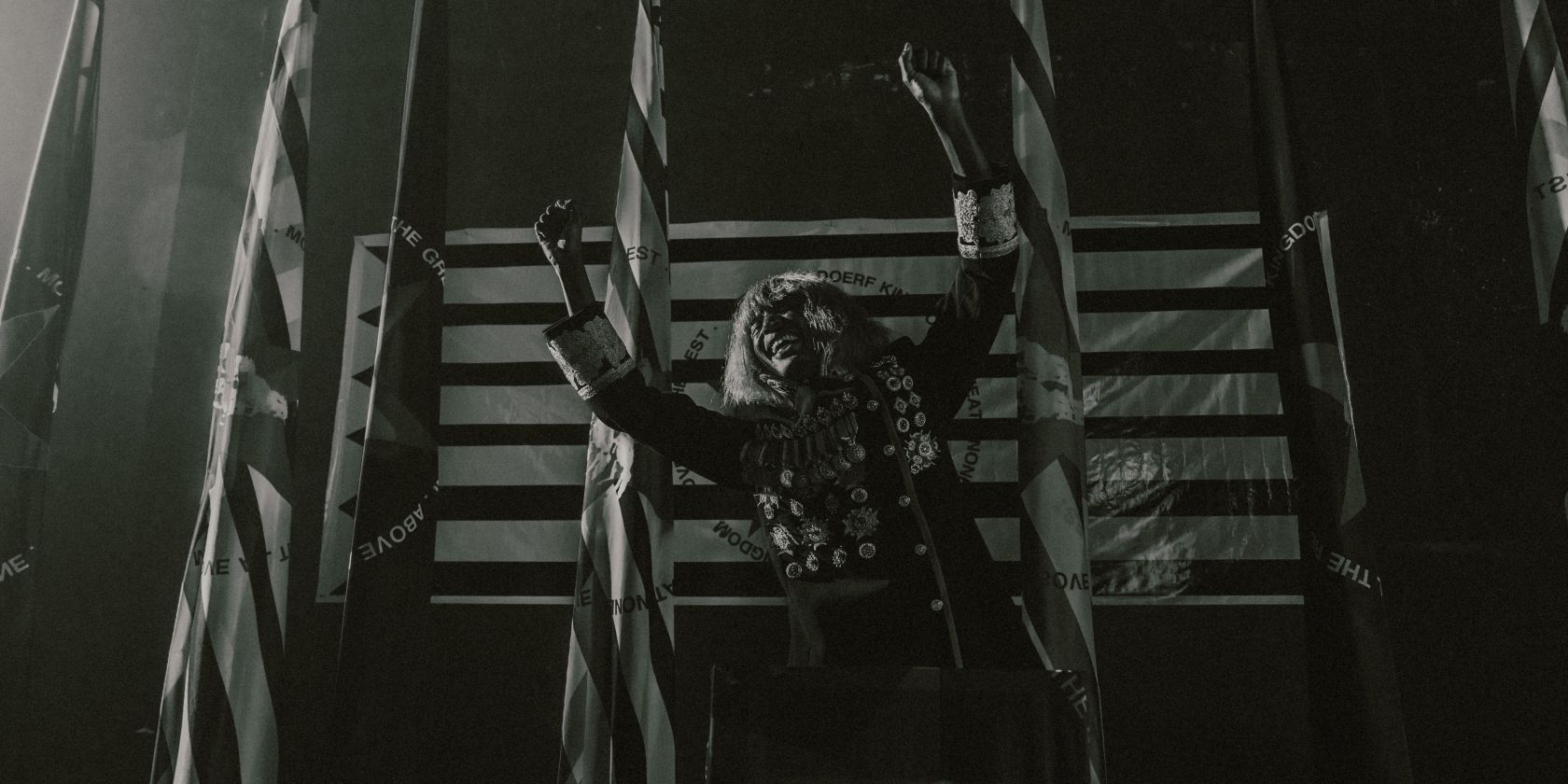 KABEAUSH&Eacute; stands on a stage in front of four flags, wearing a uniform decorated with many medals, stretching his arms sideways and laughing., &copy; Fred Odede