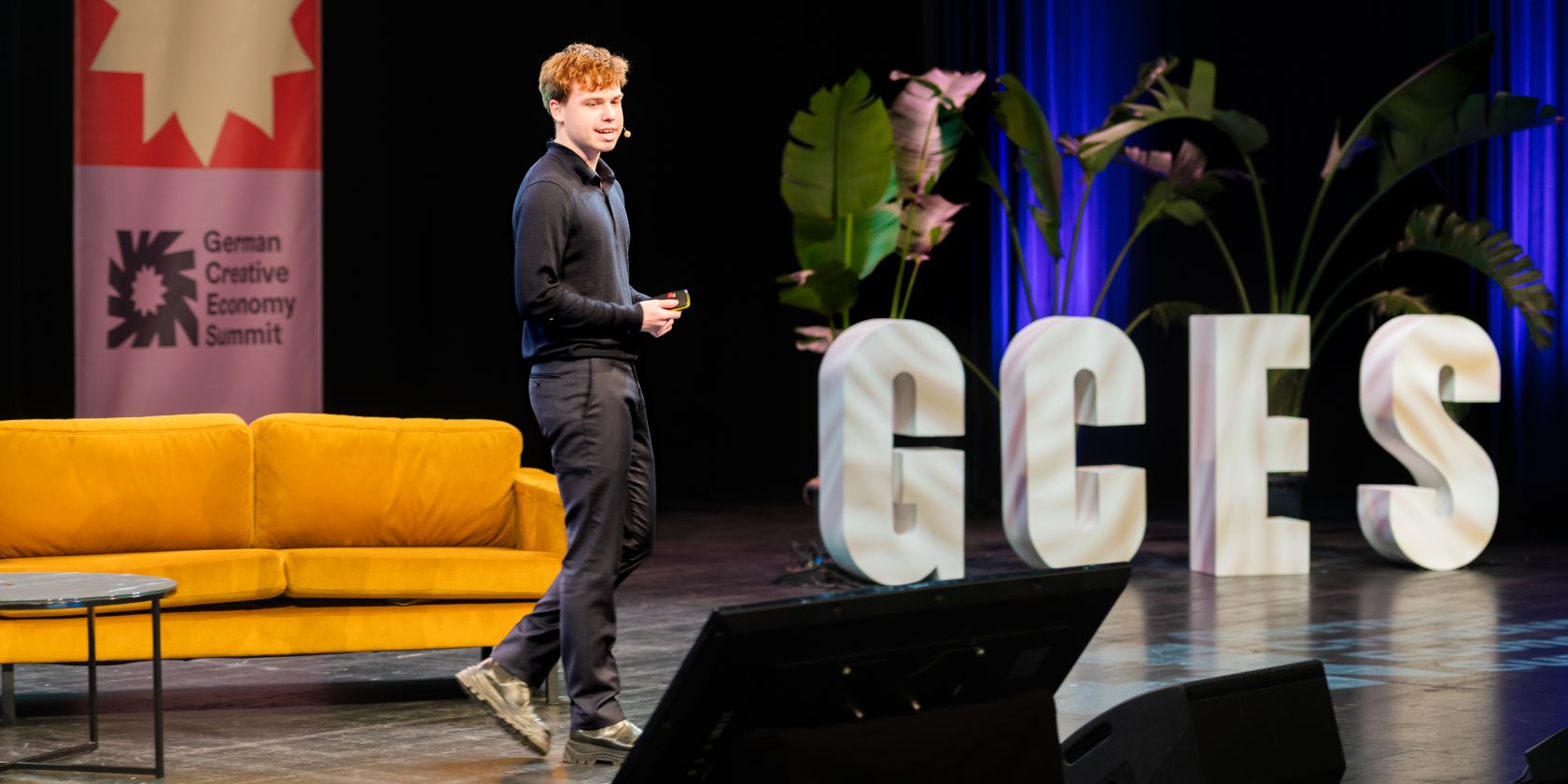 Excerpt from the German Creative Economy Summit. A stage with a speaker can be seen. In the background, there is a couch and the letters GCES., &copy; Jonas Walter