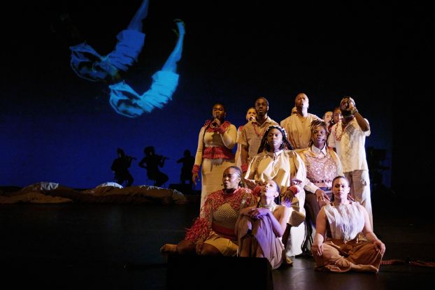 A group of performers in beige and red costumes sit and stand in formation on stage. In the background, silhouettes of musicians and a projection of a floating body are visible., &copy; Fabian Hammerl