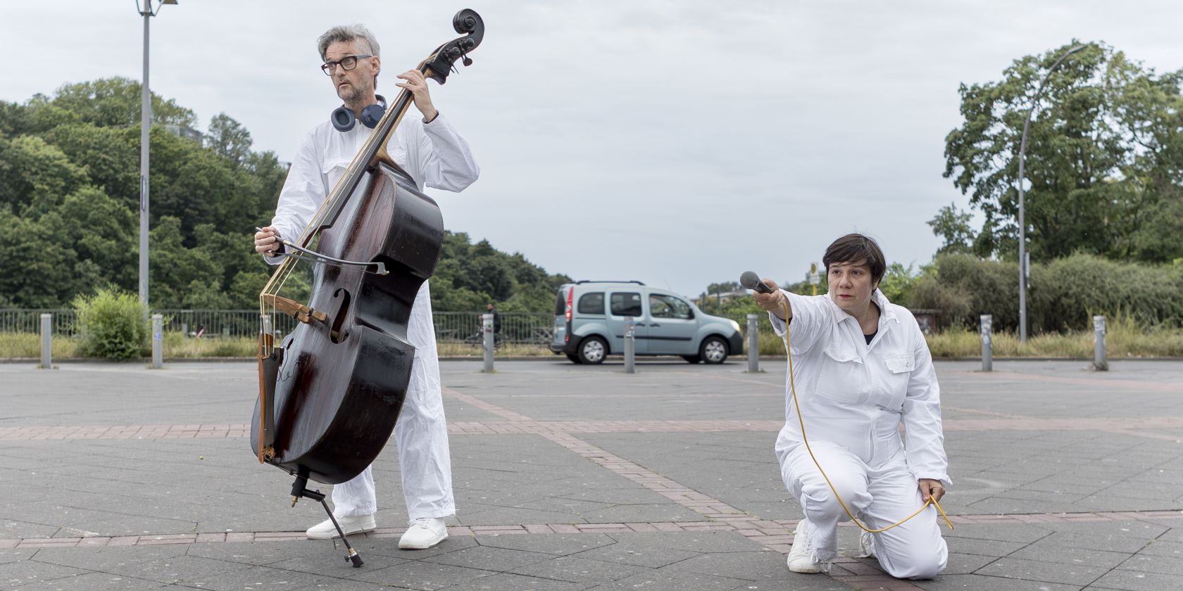 In a parking lot, there are two people wearing white full-body suits and shoes. One person is playing the double bass, while the other is kneeling and holding out a microphone with one hand., © Merlin Nadj-Torma