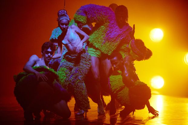 A scene from the production TRAPICANA. Several performers stand close together in a dance pose, bathed in orange light., &copy; Fabian Hammerl