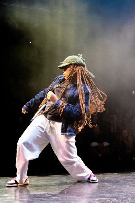 A person wearing a knitted hat, long braids, and loose clothing stands on a stage and moves their hips in one direction with bent knees. They pull themselves up in the same direction with the opposite arm., &copy; @mer.c_artistry
