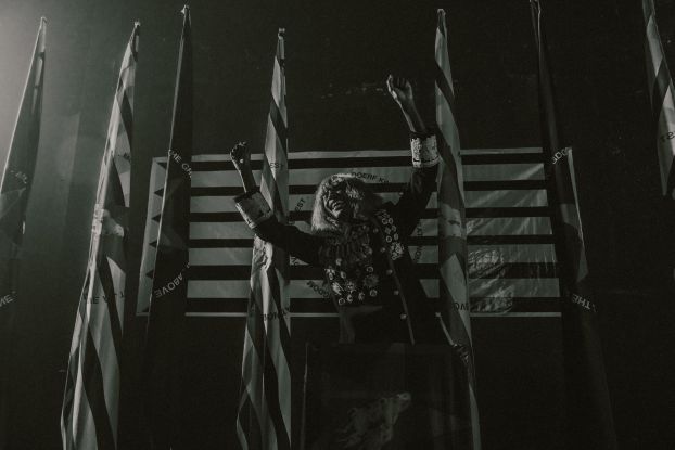 KABEAUSH&Eacute; stands on a stage in front of four flags, wearing a uniform decorated with many medals, stretching his arms sideways and laughing., &copy; Fred Odede