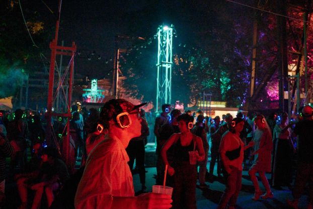 A nighttime JAJAJA event with a crowd wearing glowing red and blue silent disco headphones. In the foreground, a person wearing a cap and square glasses, holding a drink. In the background is an illuminated steel tower., &copy; Fabian Hammerl