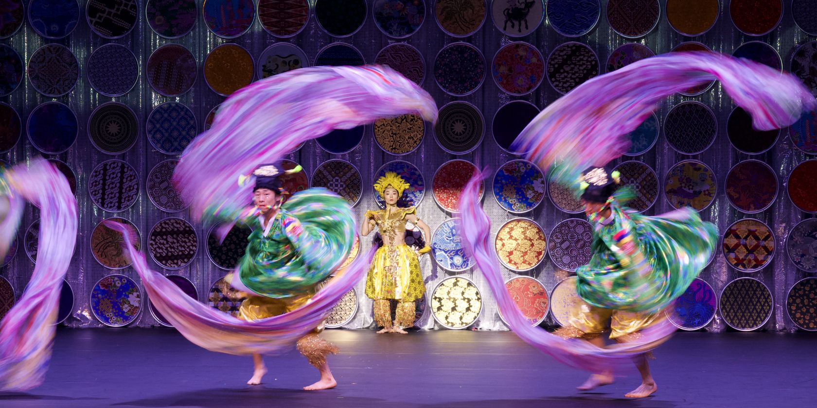 Two dancers in colourful costumes whirl bright ribbons, with one person in gold costume standing behind them in front of circular ornaments., &copy; Jean-Marie Chabot