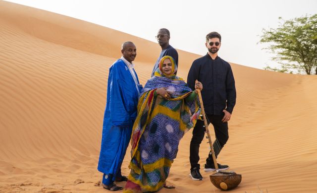 Four people are standing in front of a large dune. The person in front is wrapped in a long cloth; only their face and hands are visible. They are holding a stringed instrument in their right hand: An ardine – a Mauritanian harp., © Malika Diagana