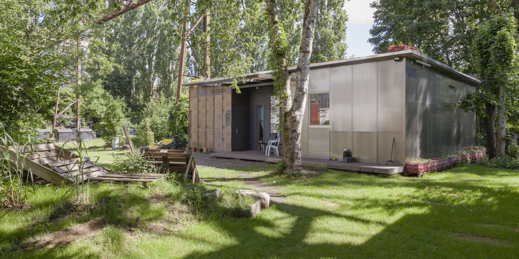 Photo of the Migrantpolitan at Kampnagel. A small one-storey house with a green garden, benches made of wooden pallets, surrounded by many trees., © L. Volkmann