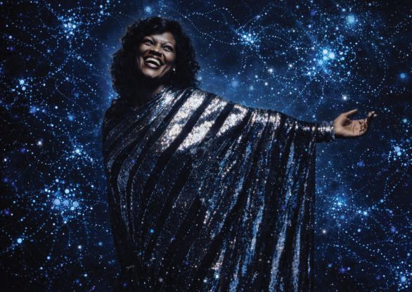 A woman stands in a glittering silver dress against the backdrop of a dark blue surface covered with many lights., &copy; Saeleen Bouvar