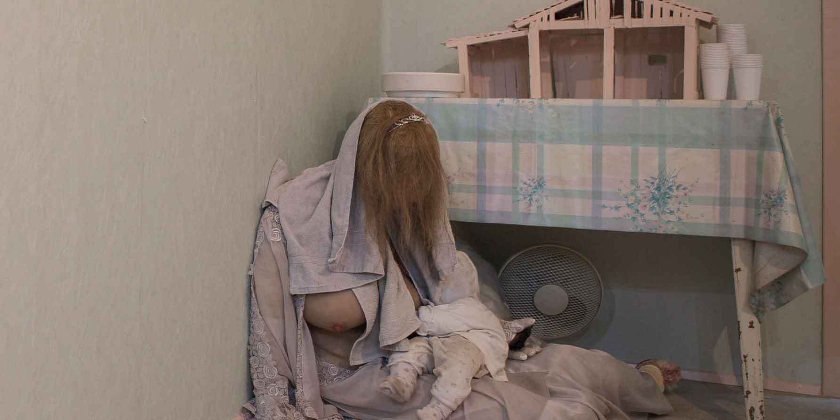 A person with long hair is sitting on the floor with a baby next to a table with a model house on it., &copy; Erich Goldmann