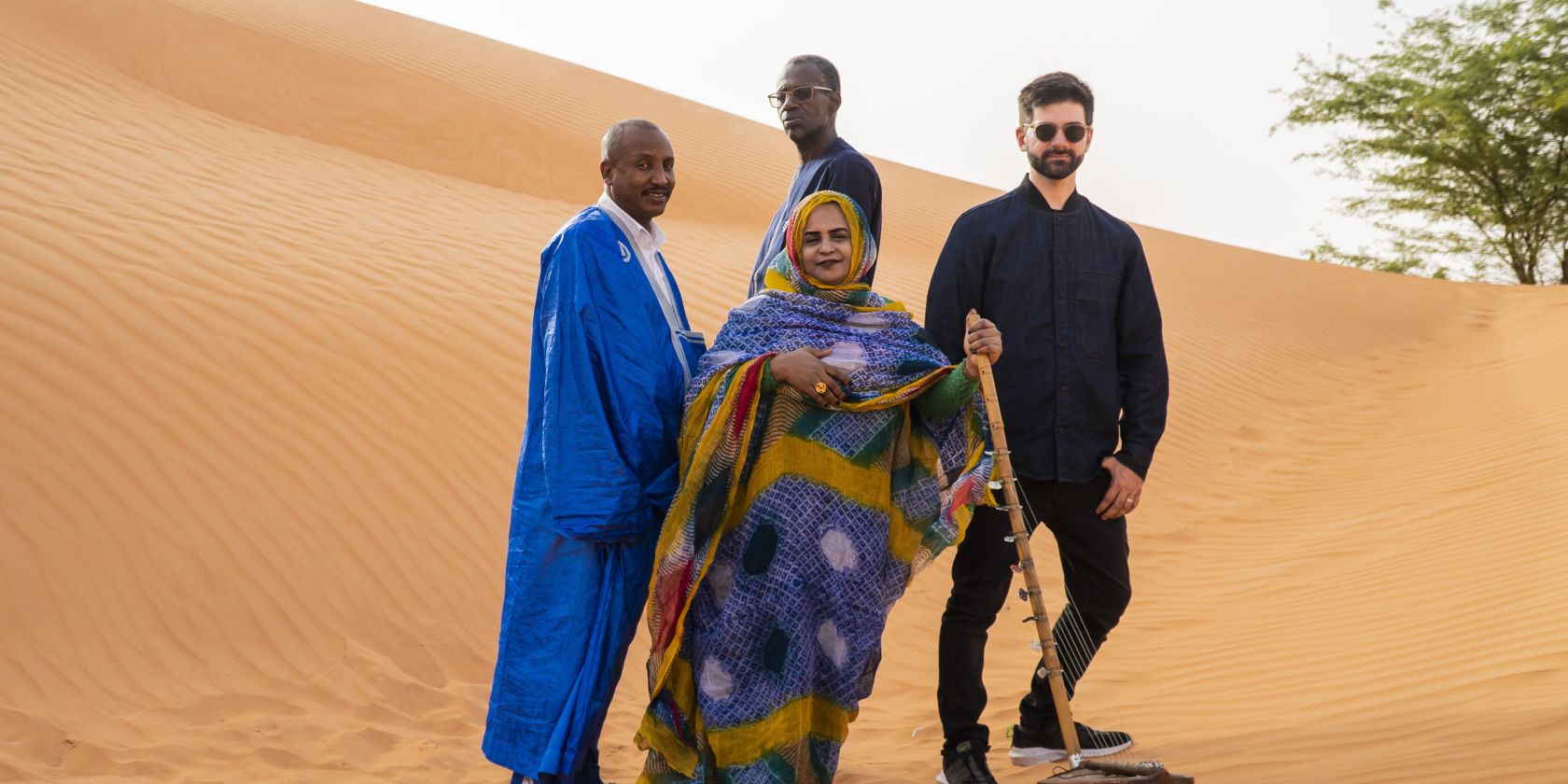 Four people are standing in front of a large dune. The person in front is wrapped in a long cloth; only their face and hands are visible. They are holding a stringed instrument in their right hand: An ardine – a Mauritanian harp., © Malika Diagana