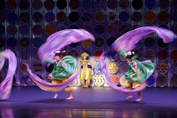 Two dancers in colourful costumes whirl bright ribbons, with one person in gold costume standing behind them in front of circular ornaments., &copy; Jean-Marie Chabot