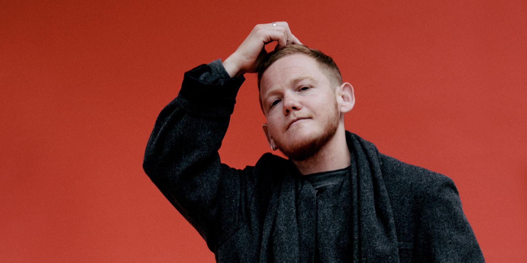 Kae Tempest stands dressed in black in front of a colorful wall with one hand on their head., &copy; Clara Nebeling