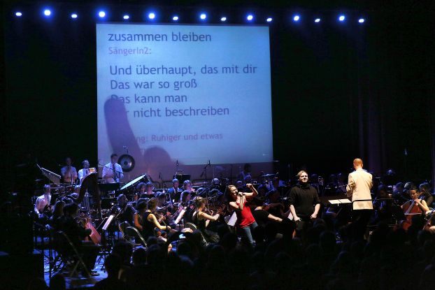 An orchestra plays on a stage, in the background is a large screen on which a song text is projected. A young woman stands in front of the orchestra, happily singing into a microphone., © Anja Beutler