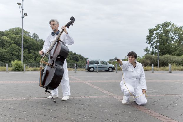 In a parking lot, there are two people wearing white full-body suits and shoes. One person is playing the double bass, while the other is kneeling and holding out a microphone with one hand., © Merlin Nadj-Torma