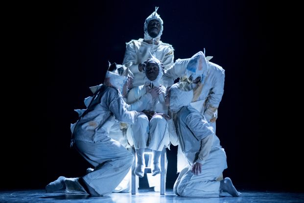 Four performers stand close to a person sitting on a chair. They are all wearing bright costumes with large spikes on the back., &copy; Camilla Greenwell