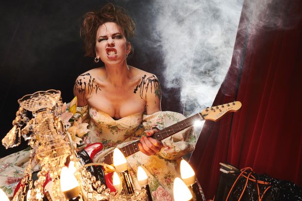 Emma Frankland, a female-presenting person with brown hair, stands in front of a curtain with an electric guitar and a rocking expression on her face., &copy; Matt Crockett