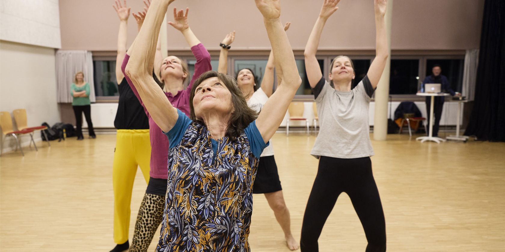 Five people are standing behind each other in a dance studio, each with both arms stretched toward the ceiling. They are wearing colorful sportswear and their eyes are directed toward the ceiling., &copy; Julie Nagel