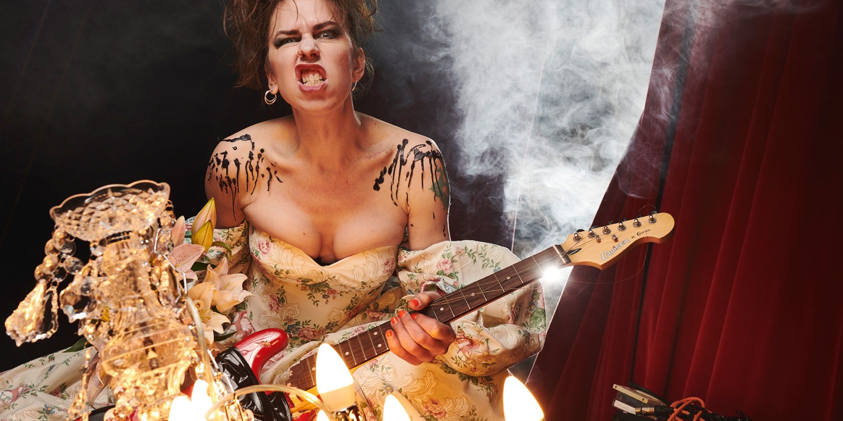 Emma Frankland, a female-presenting person with brown hair, stands in front of a curtain with an electric guitar and a rocking expression on her face., &copy; Matt Crockett