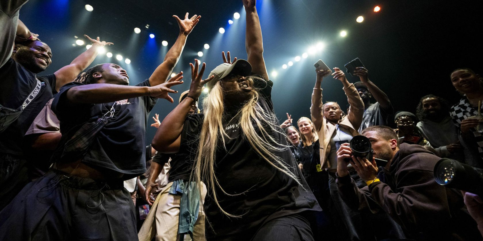 A dynamic scene in which a crowd of people stand around a dancer, cheering and cheering her on with gestures. The dancer raises her arms upwards, spreading her fingers. At the bottom right, someone photographs the dancer from close up., &copy; Teambuckmode