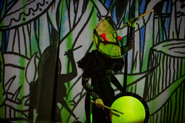 Eine Person trommelt vor einer in gr&uuml;n-schwarzen Symbolen angestrahlte Wand. Die eine Hand mit einem Drumstick in der H&ouml;he, die andere an der Trommel. Auf dem Kopf sitzt eine f&auml;cherartige Kopfbedeckung., &copy; Tsubasahori Maruszak