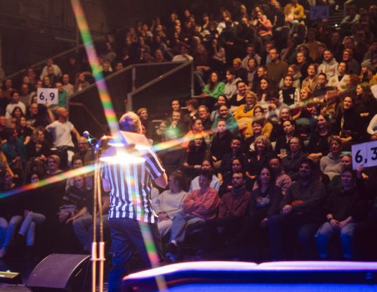 A person wearing a black and white striped T-shirt stands in front of a large audience. Two boards displaying scores are held up in the audience., © Michelle Jekel