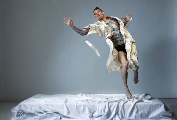 A performer in a cream silk robe and black underwear leaps from a white bed, his torso intricately tattooed., © Hugo Glendinning