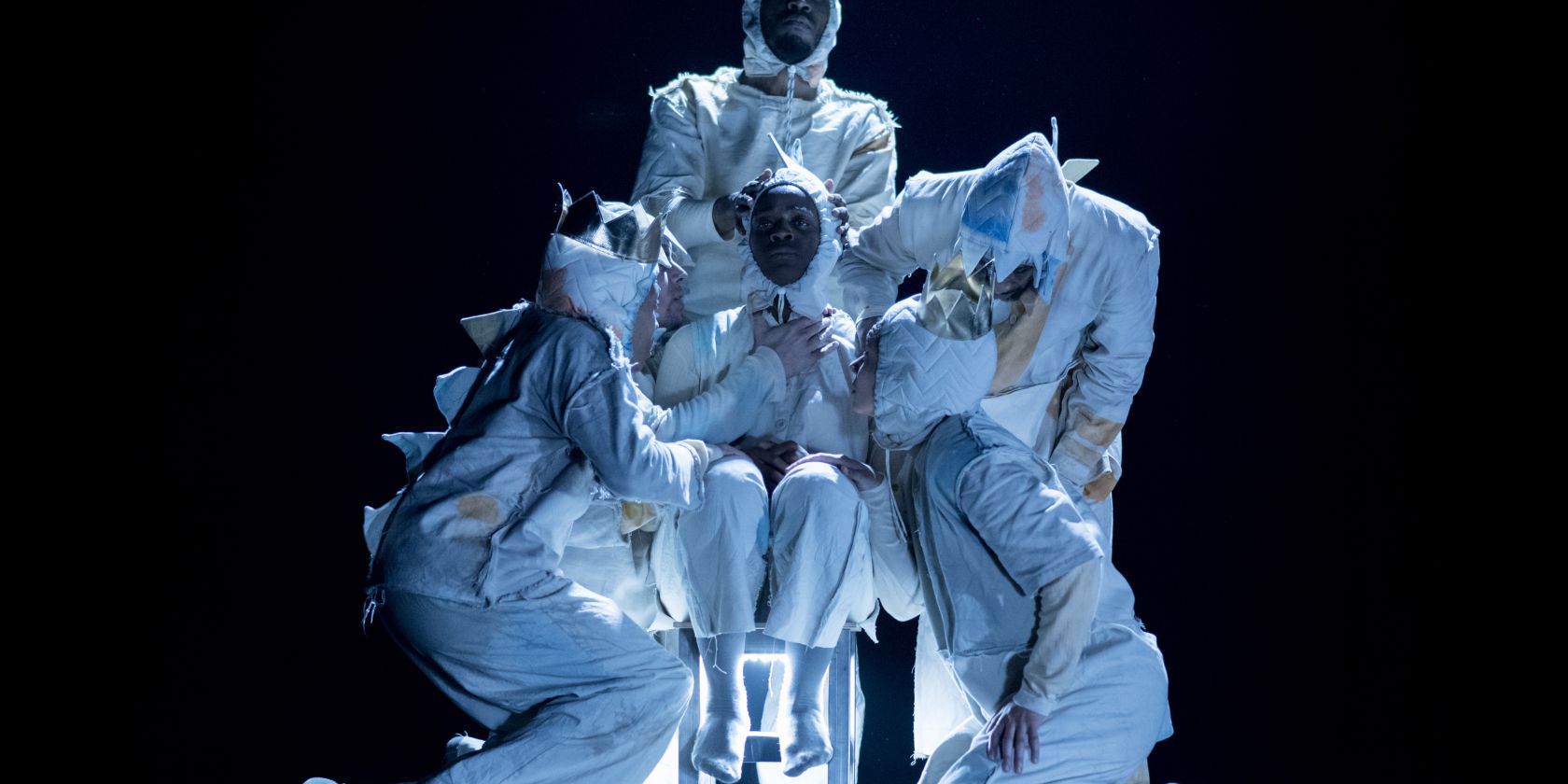 Four performers stand close to a person sitting on a chair. They are all wearing bright costumes with large spikes on the back., &copy; Camilla Greenwell