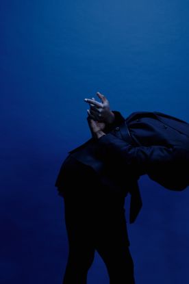 A performer is seen from the side, leaning forward, a black hoodie pulled over their head. Their hands are behind their back and stretched out., &copy; Anne Schwarzelt