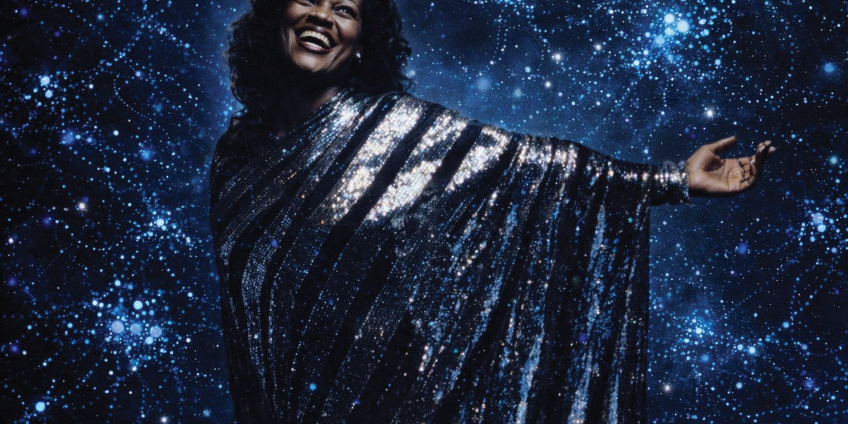 A woman stands in a glittering silver dress against the backdrop of a dark blue surface covered with many lights., &copy; Saeleen Bouvar