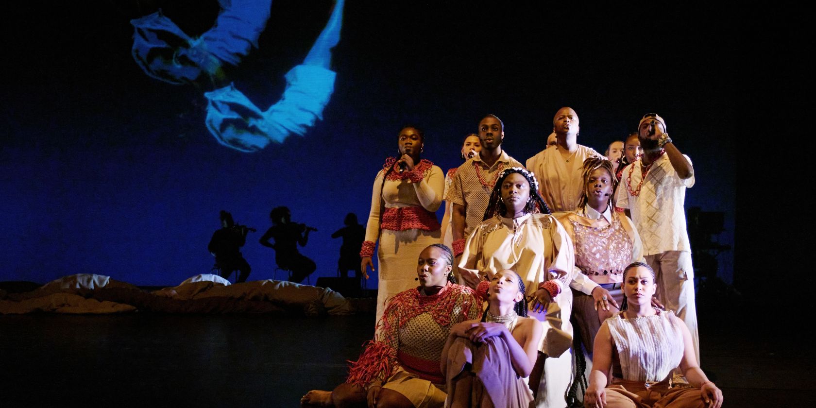 A group of performers in beige and red costumes sit and stand in formation on stage. In the background, silhouettes of musicians and a projection of a floating body are visible., &copy; Fabian Hammerl