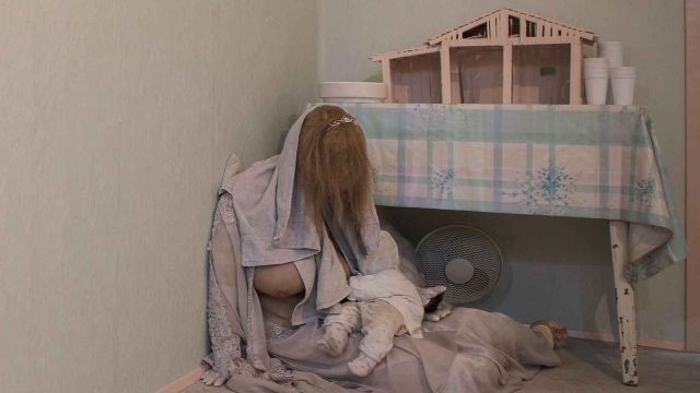 A person with long hair is sitting on the floor with a baby next to a table with a model house on it., &copy; Erich Goldmann